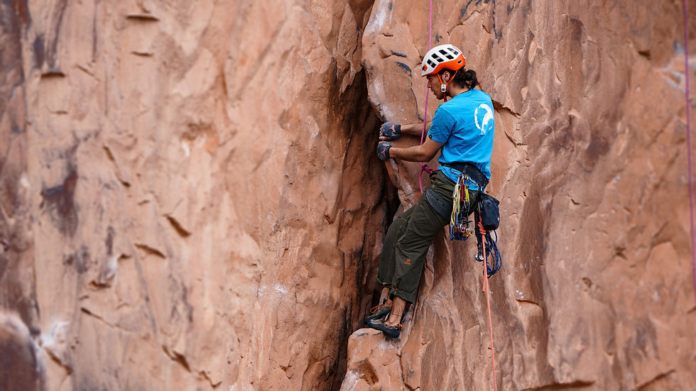 Rock climber in a blue shirt and helmet scales a rugged orange cliff face. Equipped with gear, focused on an upward climb.
