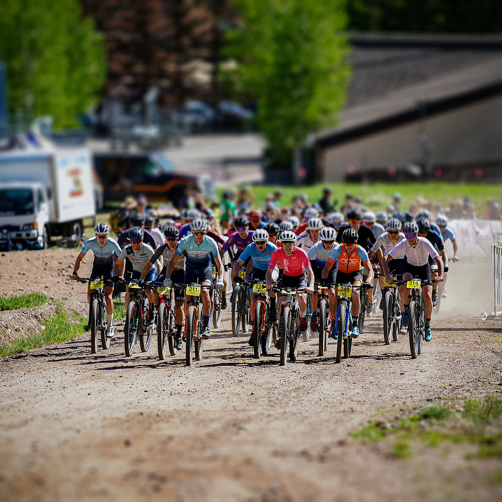 Cyclists in colorful gear race on a dirt path, surrounded by greenery. Bib numbers are visible, creating a competitive, energetic atmosphere.
