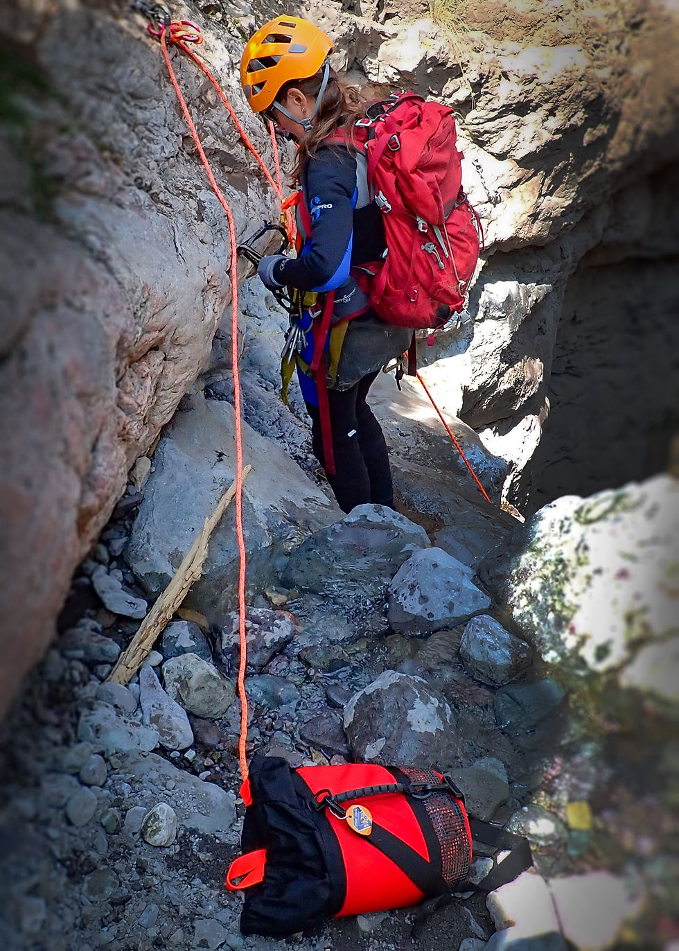 Climber in orange helmet and red backpack secures rope on rocky terrain. Bright orange rope and gear bag. Focused and determined mood.