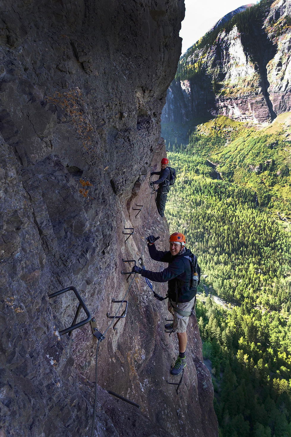 Two climbers in helmets scale steep rock with metal rungs, against a backdrop of lush green forest and mountains. Bright, sunny day.