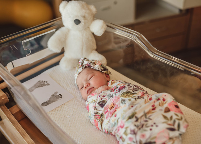 Newborn baby girl wrapped in floral swaddle. Baby is in hospital bassinet. White teddy bear behind her. 