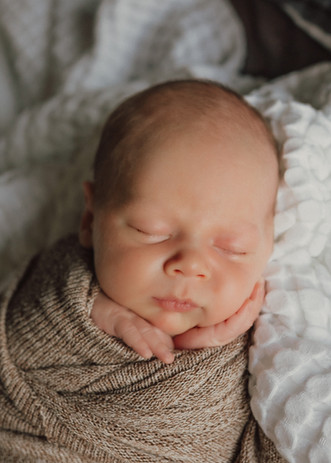 Newborn baby boy on bed. Head in hands and wrapped in tan swaddle. 