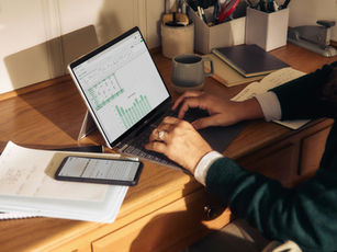 Person types at a home desk with a laptop showing a spreadsheet and bar chart, plus a phone, notebook, and coffee mug in warm sunlight.