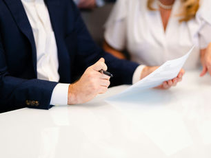 Man in navy suit reviews a document with a woman in a white blouse at a bright table, pen in hand, focused and professional.