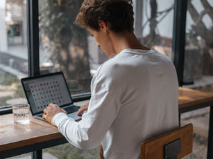 Person in a white shirt works on a laptop at a café table beside a glass of water, seen from behind.