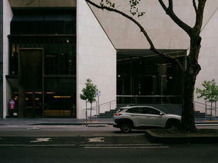White SUV drives past a modern beige building with dark glass, street trees.