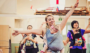 A mother with blonde hair carrying her 8 month old baby in a blue woven wrap is frozen in a dance move sliding to the side arms high. Two mothers are in the background also carrying their babies in structured baby carriers. The babies are all facing towards their mothers. The mothers are all smiling.