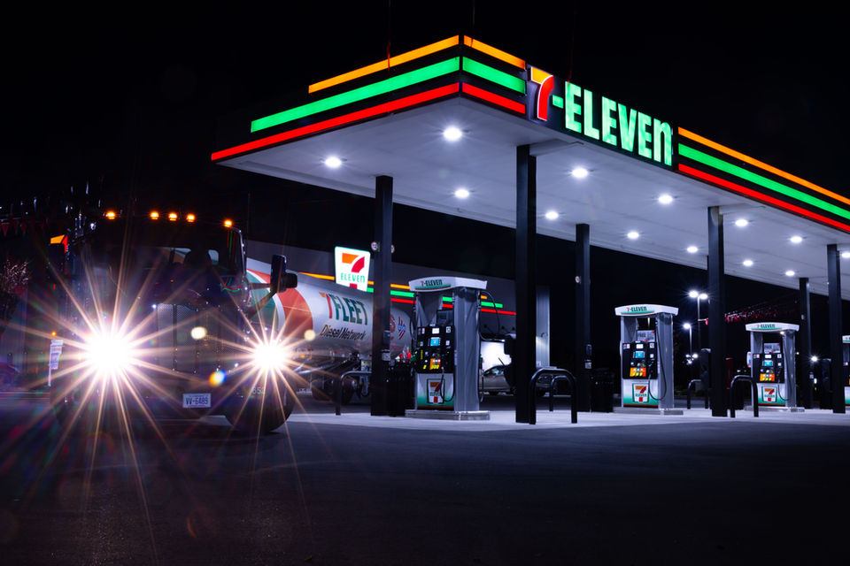 Close shot of 7 Fleet truck with headlights creating starbursts, approaching camera under 7-Eleven canopy lights at night.