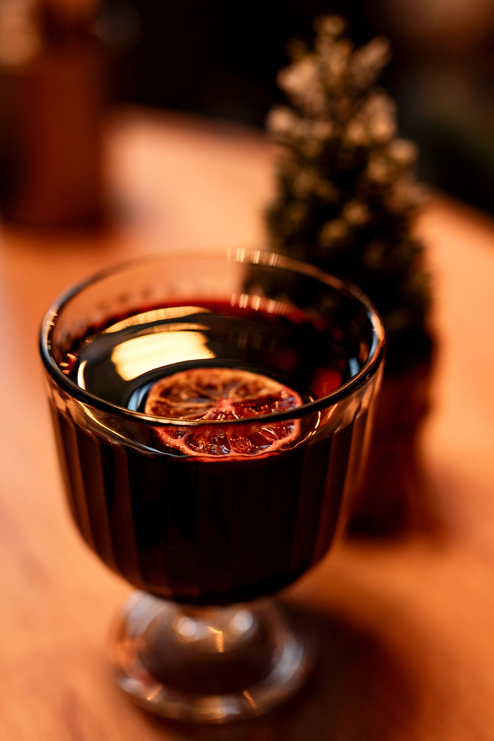 高脚杯中盛有热红酒,漂浮着一片橙片。背景模糊,灯光温暖,营造舒适节日氛围。