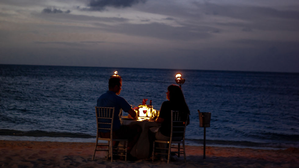 A couple dines at a candlelit table on the beach at dusk, with the ocean as a backdrop. Two lit torches create a warm, romantic ambiance.