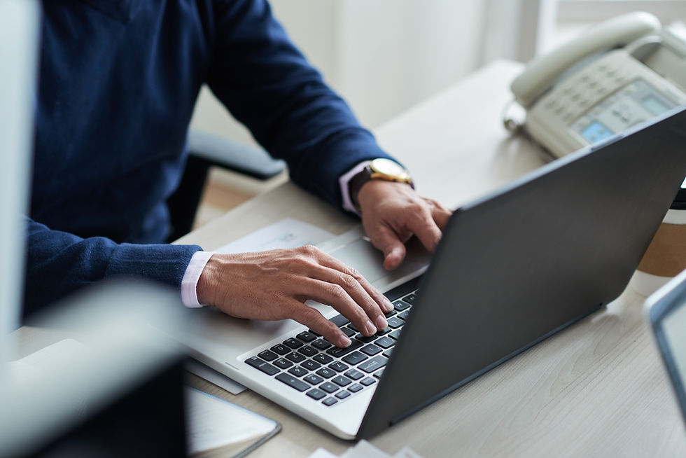 A close-up view of a person's hands typing on a laptop in an office environment. The desk includes a phone, a cup, and other office supplies, indicating a productive workspace. The person appears focused on their work.