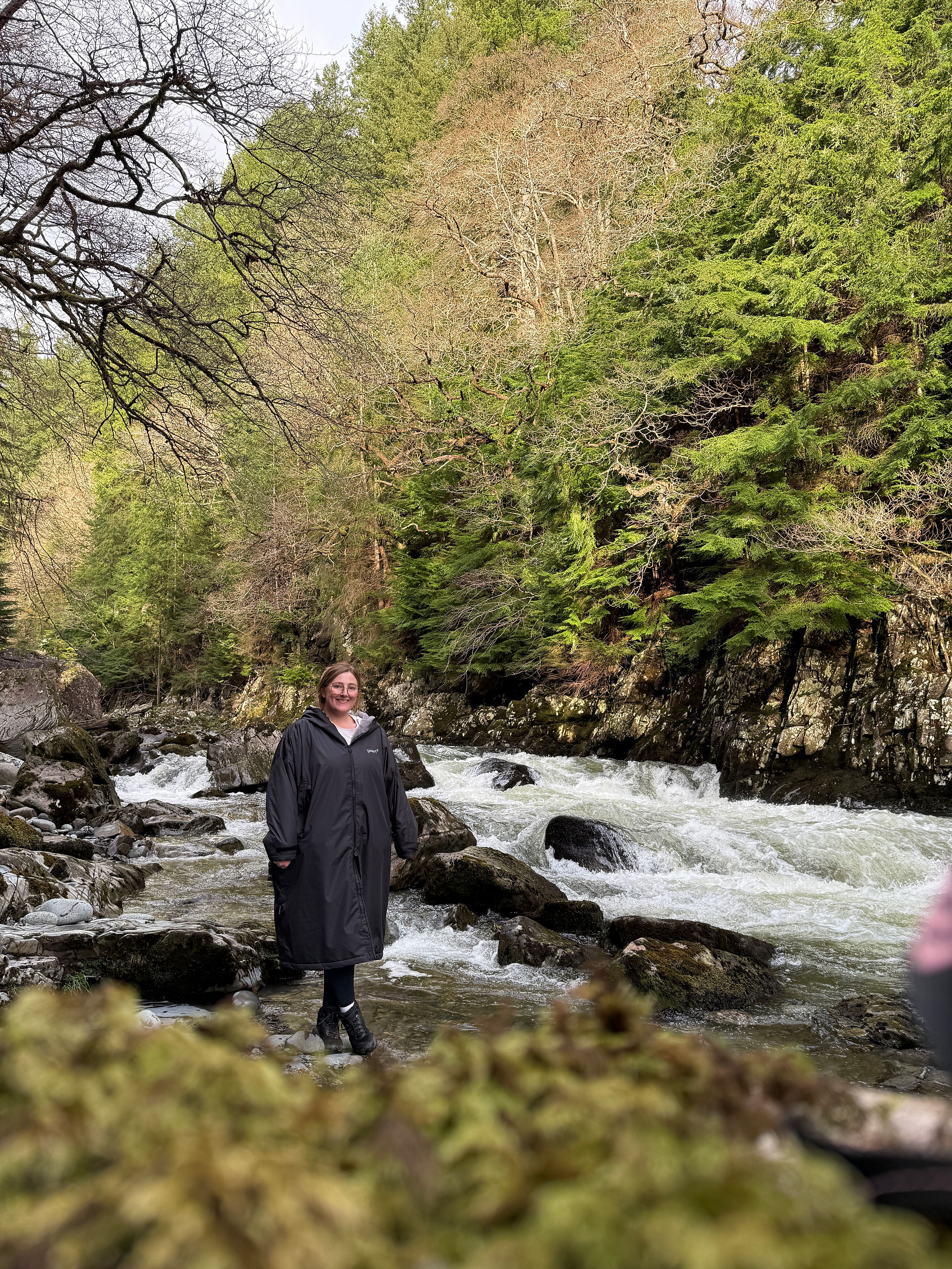 Chloe in a large black coat, smiling at the camera from a distance. She is walking next to a river. There is a big hill behind her. Lots of greenery.
