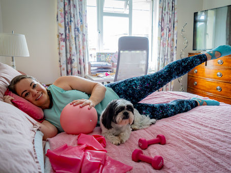 Leanne lying on her side on her bed with her leg raised, resting a hand on a pink ball. Stanley her dog, pink rsistance band and pink weights are in front of her. She is wearing blue leggings and short sleeved top.