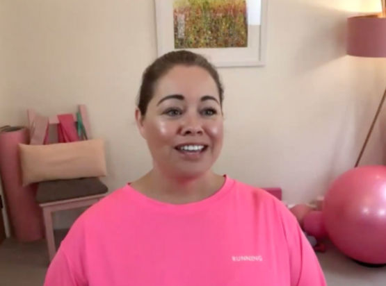 Leanne speaking to camera in a neon pink top with pink fitness equipment in the background.