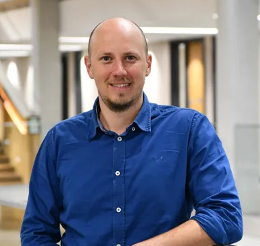 Bald man in a blue shirt smiles in a modern indoor setting with soft lighting and a staircase in the background. Warm, welcoming mood.