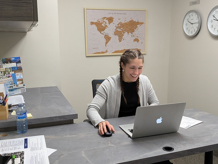 CPG's Jessica working on a laptop in an office. Map and clocks on wall. Gray desk with papers and a water bottle.