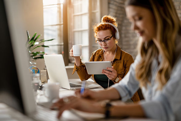young-creative-businesswoman-wearing-headphones-while-surfing-net-digital-tablet-drinking-