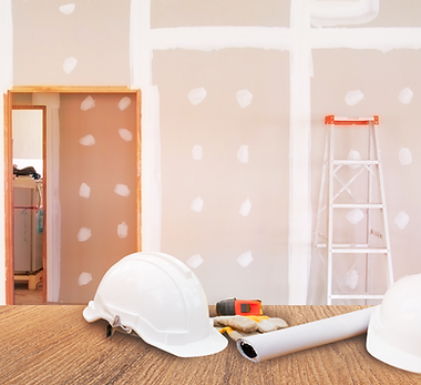 Drywall installation project with exposed joint compound, hard hats, blueprints, and construction tools on a wooden table.