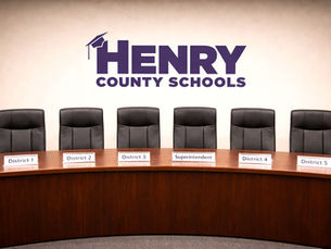 Empty Henry County Schools boardroom with a curved wooden conference table and six black leather chairs. Nameplates in front of each seat read District 1, District 2, District 3, Superintendent (center), District 4, and District 5. The Henry County Schools logo is displayed prominently on the wall behind the table.