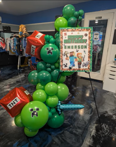 Close-up of towering Minecraft-themed balloon centerpiece for Emerson's birthday, featuring red TNT cube balloons stacked high, green Creeper-inspired accents, a large black '6' balloon, diamond sword foil balloon, custom pixel art sign, and base of black and green balloon clusters on a dark backdrop.