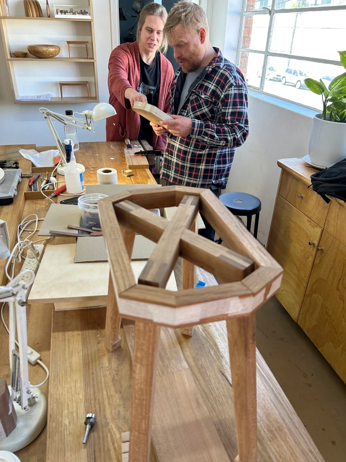 Students discussing stool components and joinery at the workbench.