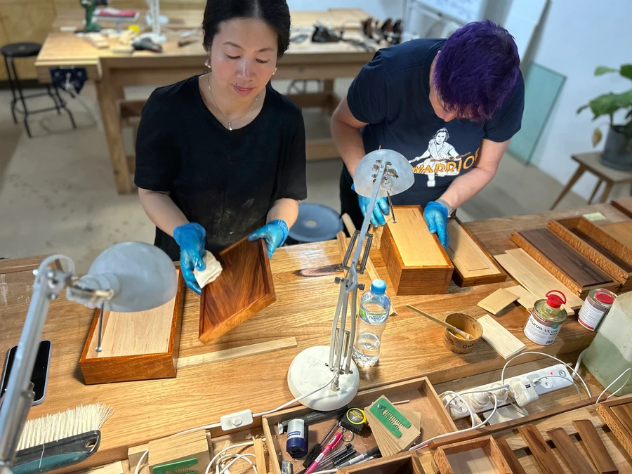 Two students applying finish to timber keepsake boxes at a workbench.