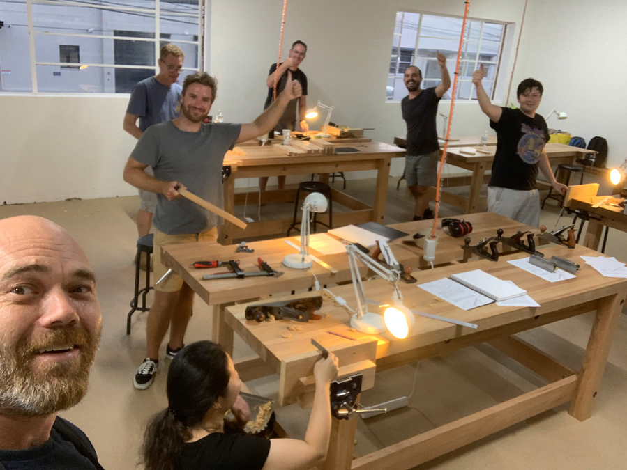 A class group smiling and giving thumbs-up around the workshop benches.