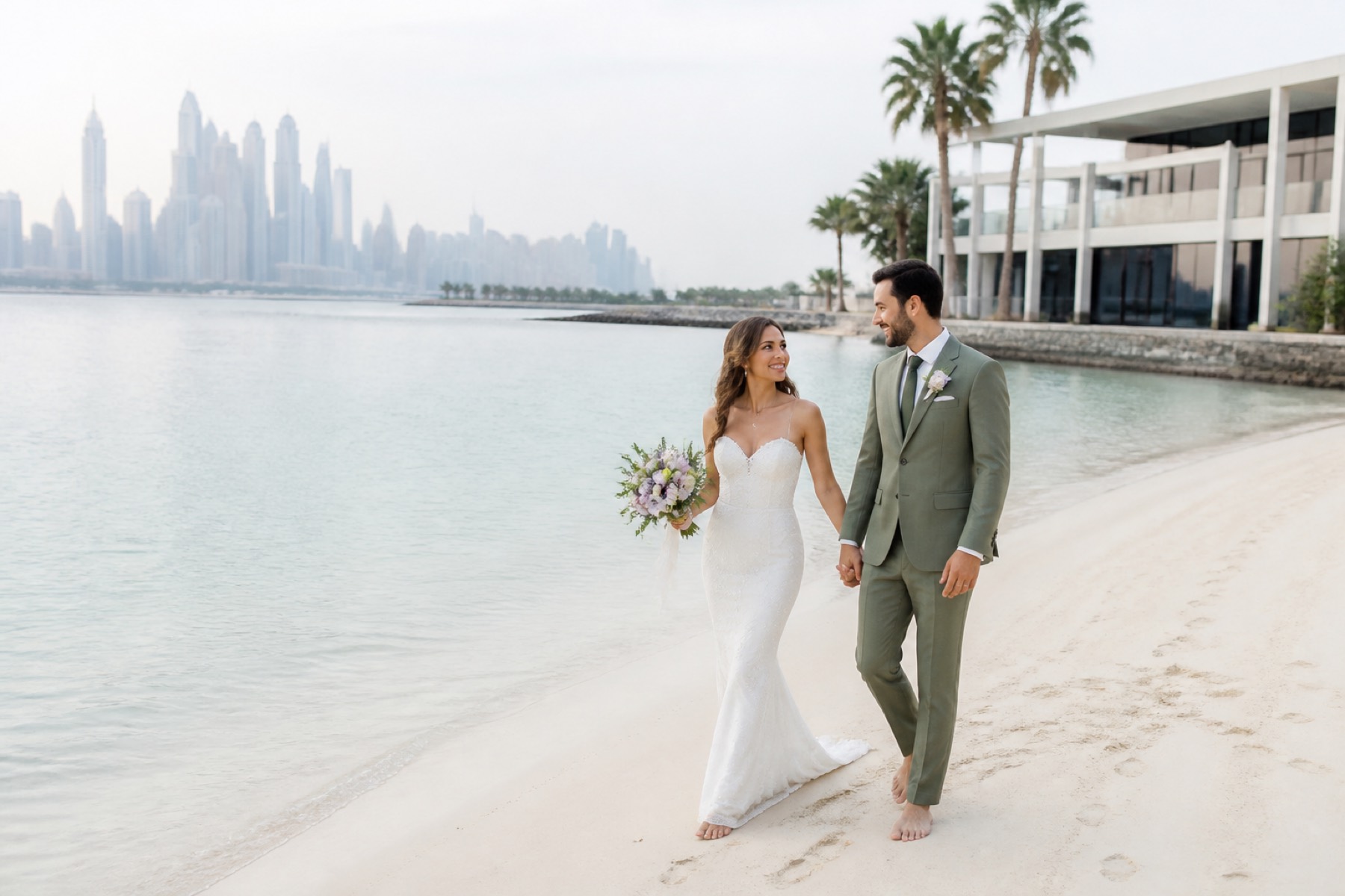 Bright shoreline at a private Dubai resort with a couple walking beside the water and skyline in distance; no arch or décor.