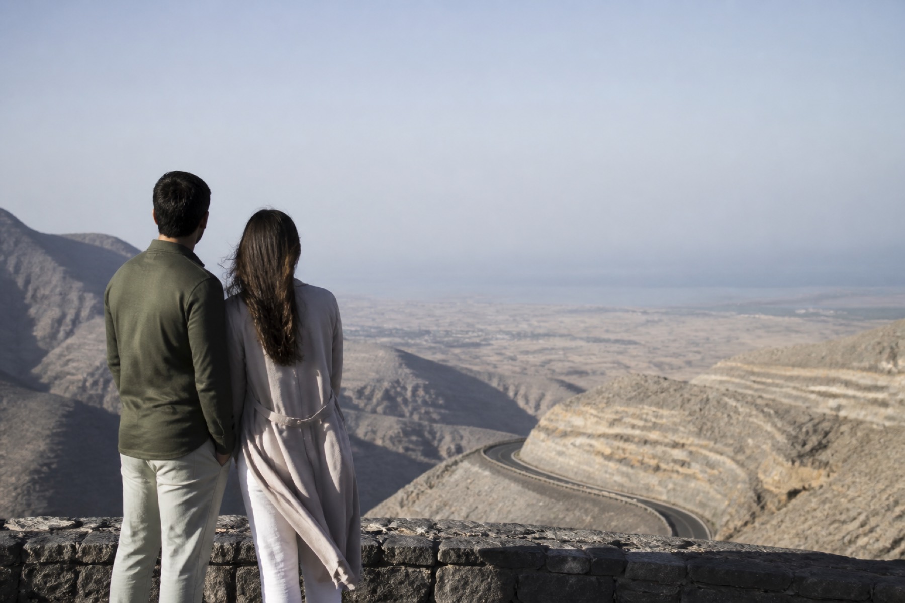 Cool daylight view from Jebel Jais toward desert and sea with a sophisticated couple at a modern lookout; no décor.
