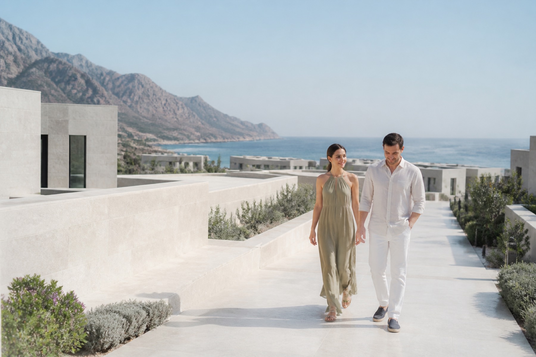 Bright Hajar Mountains meeting the blue Gulf of Oman, with a couple walking through a modern resort landscape; no décor.