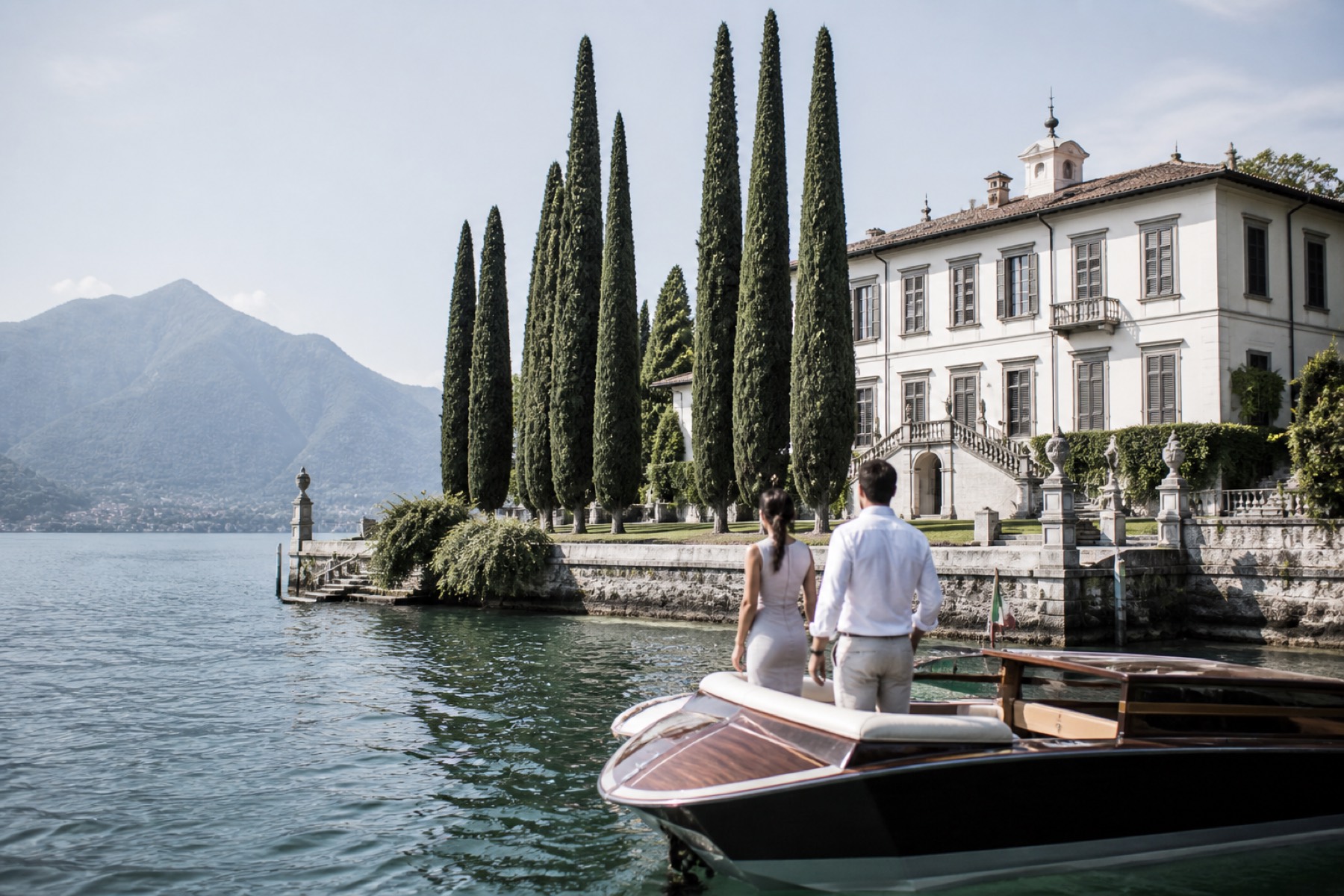 Bright Italian lakeside villa and cypress garden with a couple arriving by boat; minimal setting, no décor.