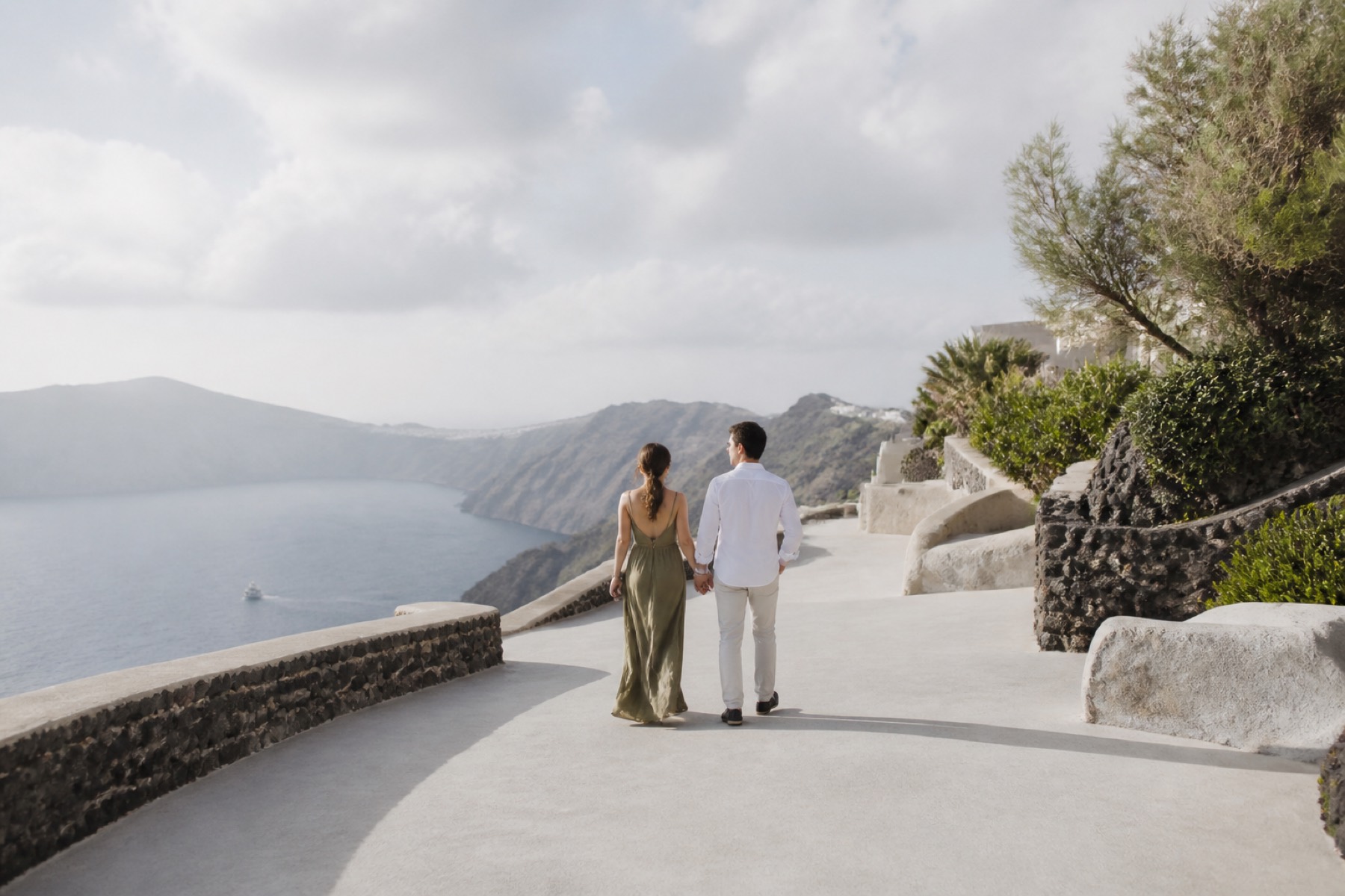 Bright caldera view from pale volcanic-stone architecture with a couple walking at blue hour daylight; no décor.