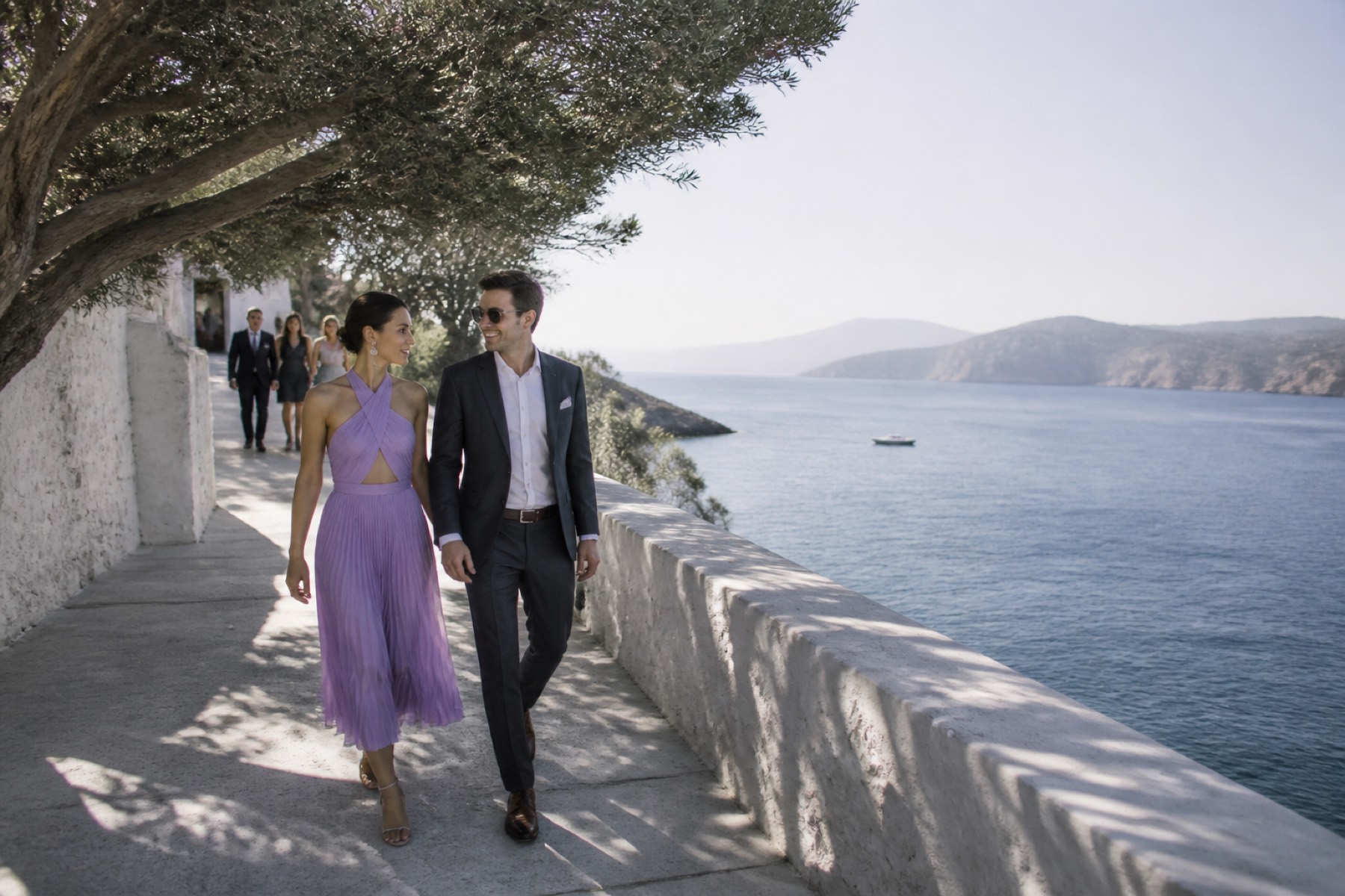 Bright Aegean sea and restrained white stone architecture with guests arriving along a shaded walkway; no décor.