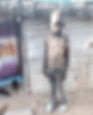 Young boy in worn clothes stands outside a corrugated metal hut with a thatched roof. Purple curtain and black sandals are visible.