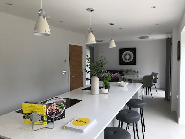 Modern kitchen with a white island, bar stools, pendant lights, and plants. Cookbooks are open on the counter. Simple, bright ambiance.