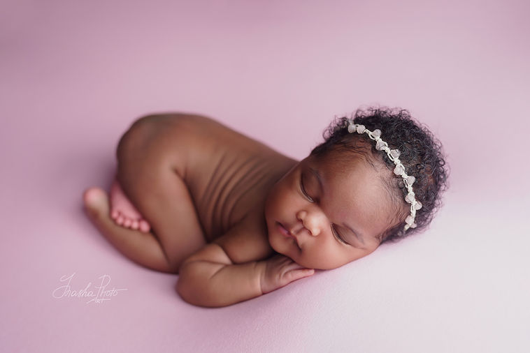 Dark skinned newborn baby girl sleeping peacefully on a pink background, wearing a pearl crown during a newborn photoshoot