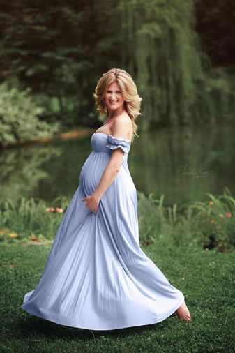 Donna incinta in abito azzurro lungo posa sorridente vicino a un laghetto durante servizio fotografico di gravidanza