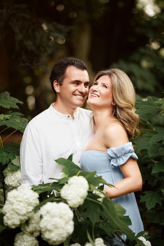 Coppia sorridente in attesa posa tra ortensie bianche, lei in abito azzurro e lui in camicia bianca, durante servizio fotografico di gravidanza all'aperto