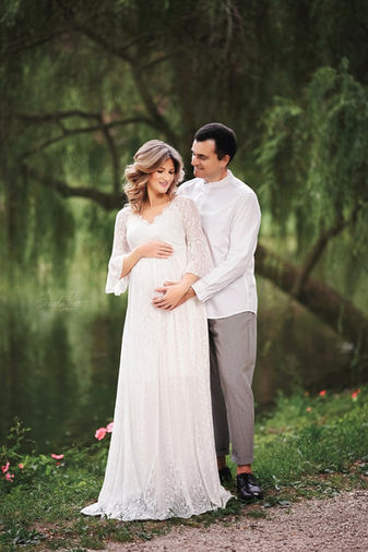 Coppia in attesa posa in un parco, lei in abito bianco lui in camicia bianca, durante servizio fotografico di gravidanza