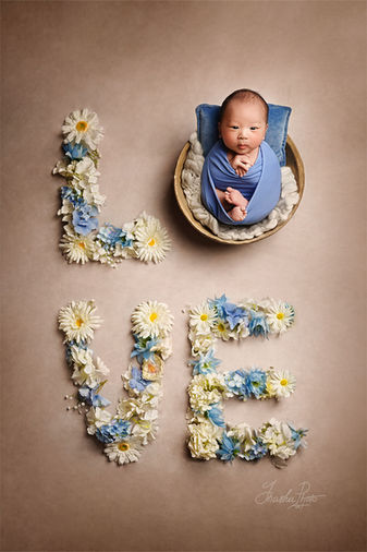 Awake newborn baby in blue fabric in a basket, surrounded by the word LOVE formed by white and blue flowers in an artistic newborn photoshoot