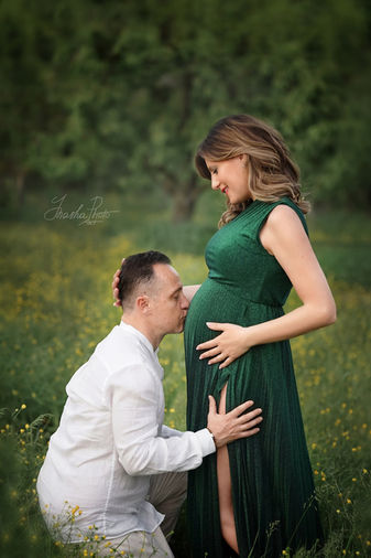 Coppia in attesa in un prato fiorito, futuro papà bacia il pancione della mamma in abito verde