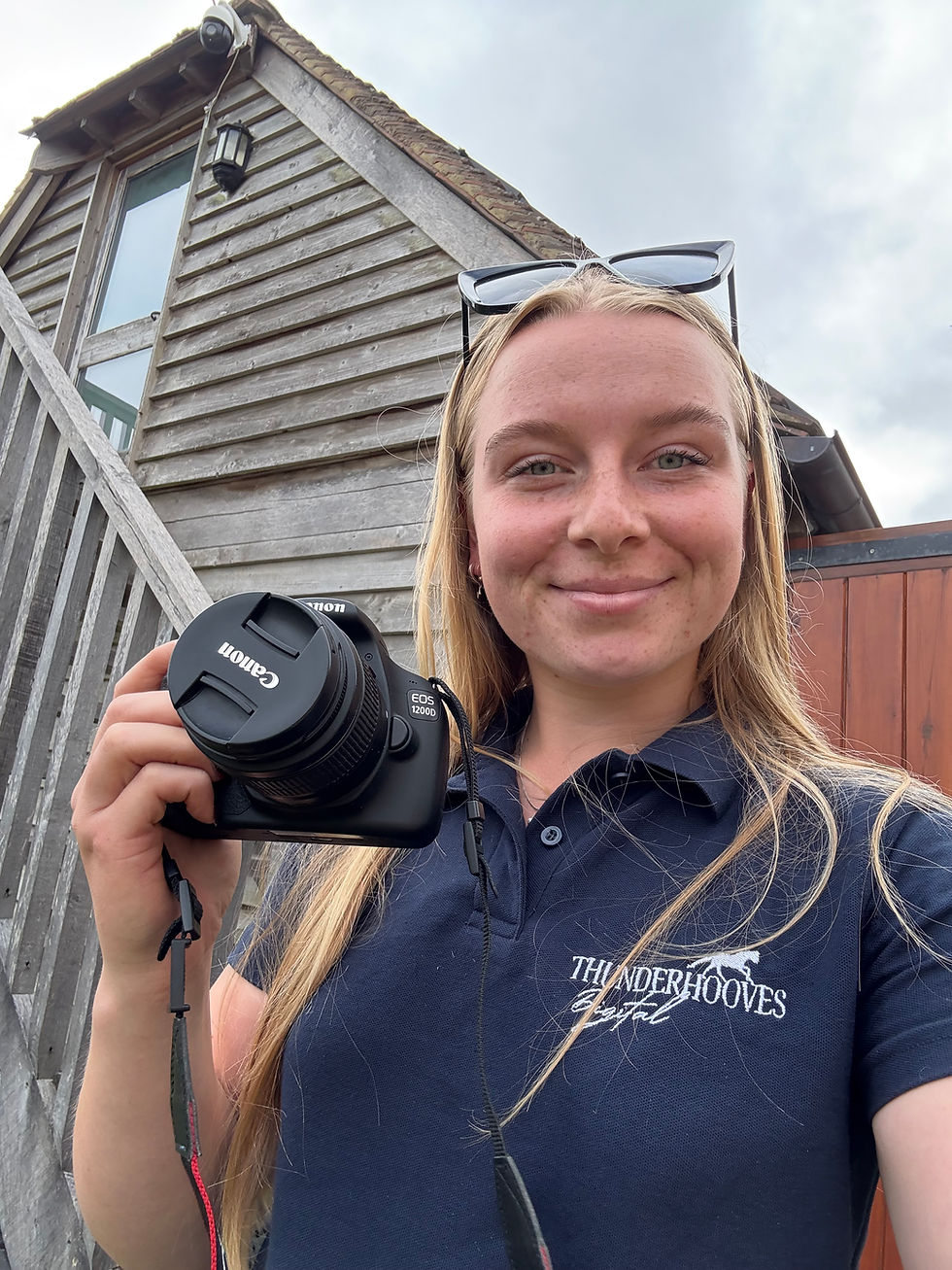 Woman smiling, holding a Canon camera. She's wearing a navy shirt with "THUNDERHOOVES Digital." Background shows a wooden building. Sky is overcast.