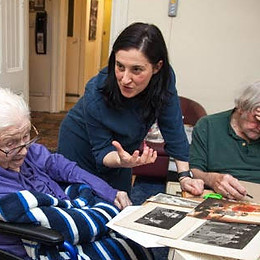 Fiona, a white woman with shoulder length dark hair, is leaning over a table and gesticulating passionately, her face mid-speech and happy. Two elderly woman sit either side of her.