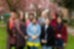 A group of eight colourfully-dressed women smile to the camera. There are blossom trees in the backg