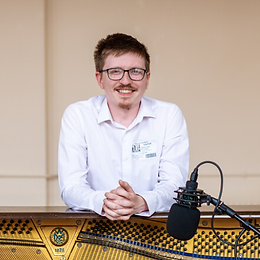 Jobe, a white man with ginger hair, a moustache and a small goatee, smiles to the camera. He is wearing a white shirt and leaning over a soundesk, behind a microphone.