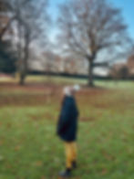 Woman in a black coat and colorful hat stands on a grassy field with fallen leaves. Bare trees in the background under a clear sky.