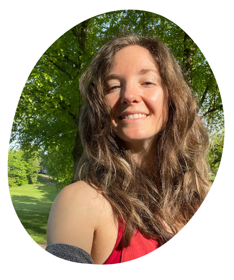 A woman smiling in the sunshine with brown wavy hair and glowing cheeks and green trees behind her