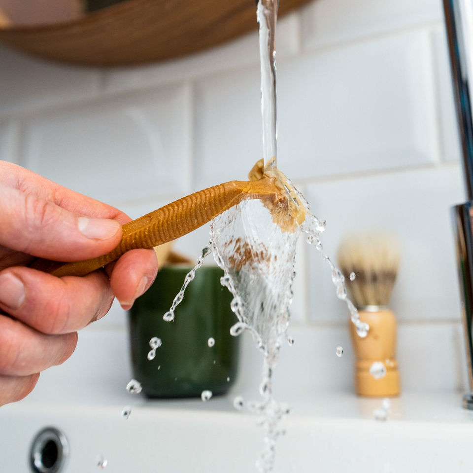 Campaign image of a bamboo razor being held under the running tab, by male hand, in a bathroom setting.