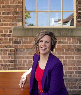 Professional headshot of a woman looking at the camera with a brick background, purple blazer and red top.