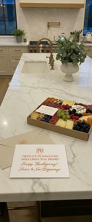 A beautifully arranged charcuterie board and a personalized "Welcome Home" card from Princeton Orchard sitting on a custom Calacatta marble kitchen island with an unlacquered brass bridge faucet following a luxury home remodel in Houston.